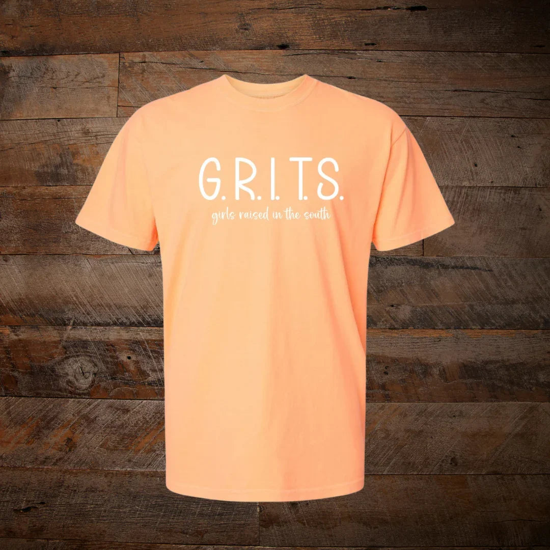 Peach t-shirt with 'G.R.I.T.S. girls raised in the south' text, rustic wood background
