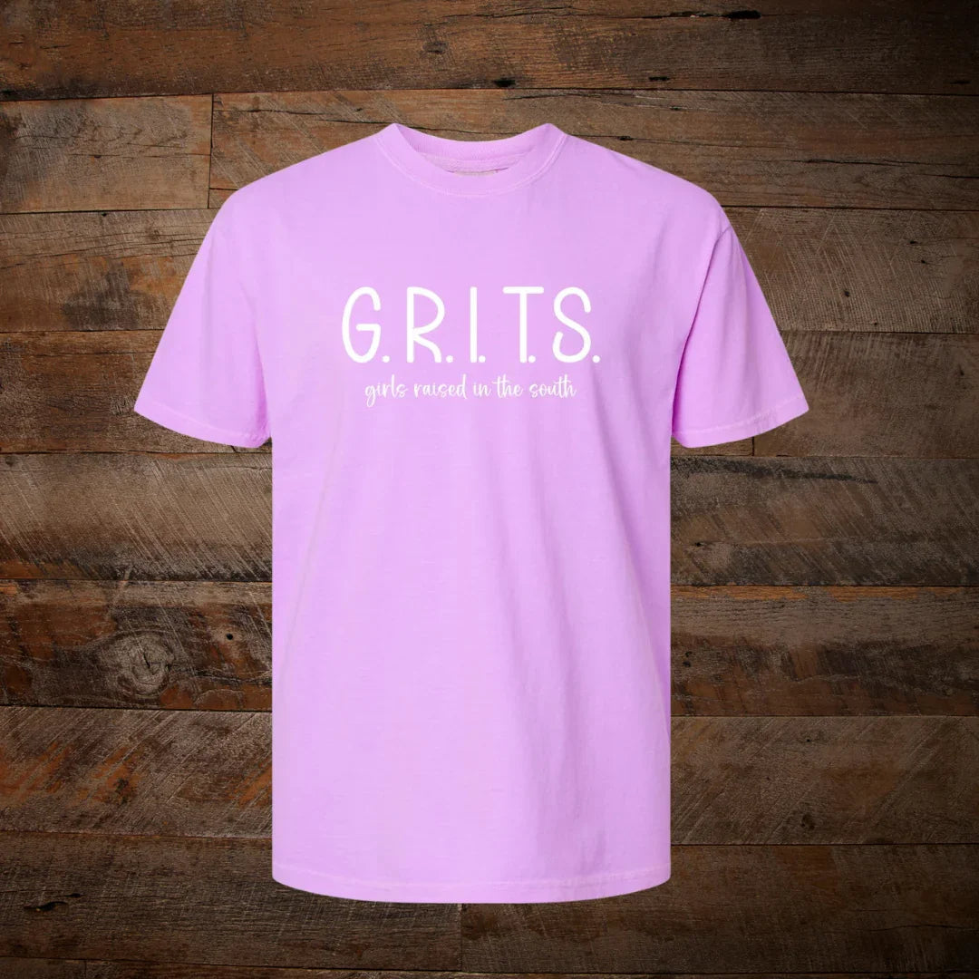 Light purple t-shirt with white text 'G.R.I.T.S. girls raised in the south' on rustic wood background