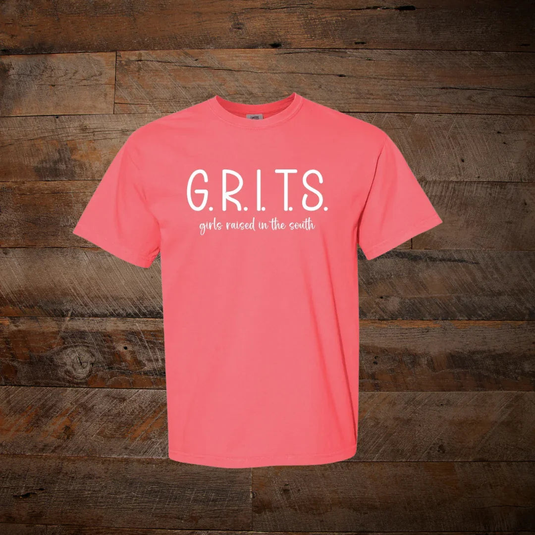 Coral pink t-shirt with 'G.R.I.T.S. girls raised in the south' text on rustic wood background.