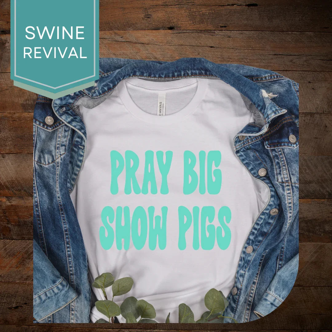 White t-shirt with teal Pray Big Show Pigs text, styled with denim jacket on wood